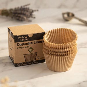 May include: A stack of three light brown, fluted paper cupcake liners next to a square cardboard box labeled Bake Choice Cupcake Liners Large Size with a graphic of a liner and the number 100, featuring a brown and black color scheme.