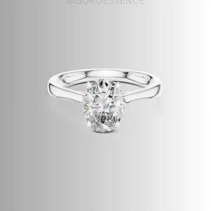May include: A silver-toned ring featuring a central oval-cut clear gemstone held by four prongs on a smooth, polished band. The text MGOROESSENCE is visible above the ring. The design is simple, focusing on the faceted stone and metal band.