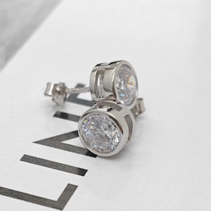 May include: A pair of silver-toned stud earrings featuring round, clear, faceted stones set in a bezel setting. The earrings have a metallic finish and are positioned on a white surface with black text that reads CELINE and CELINE.COM.