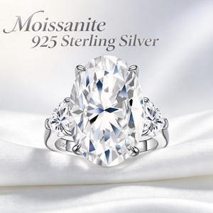 May include: An oval-cut moissanite ring set in 925 sterling silver. The ring features a large central oval stone flanked by two smaller oval stones on each side. The band is stamped with S925. Text above the ring reads Moissanite 925 Sterling Silver.