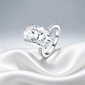 Op de afbeelding: Een zilverkleurige ring met een grote, ovaal geslepen heldere edelsteen die door vier pootjes wordt vastgehouden. De ringband is slank en metaalachtig, versierd met een rij kleine, sprankelende pavé-gezette steentjes langs het bovenste gedeelte.