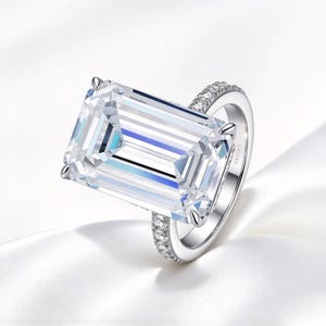 May include: A silver-toned ring featuring a large, rectangular emerald-cut clear center stone held by four prongs. The band is accented with a row of small, round pavé-set clear stones along the sides, creating a bright, reflective appearance.