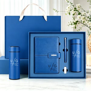 May include: A blue gift set containing a notebook, a pen, a USB drive, and two vacuum-insulated tumblers. Each item features the white printed text U/G and Uzma Ghaffar. The set is presented in a blue box with a matching blue gift bag with handles.