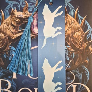 Op de afbeelding: Blauwe boekenlegger met een witte silhouet van een huilende wolf. De boekenlegger heeft een blauwe kwast. De achtergrond is een boekomslag met gouden en blauwe accenten.