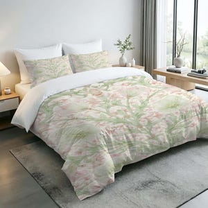 May include: A bedding set featuring a duvet cover and two matching pillowcases with a repeating botanical pattern of pale pink flowers and light green stems on a white background. The set is displayed on a bed with two solid white pillows.
