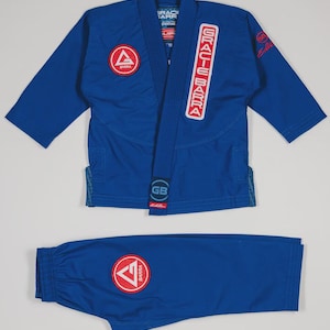 May include: A blue Jiu-Jitsu uniform set, comprising a jacket and trousers. The jacket has a red and white logo patch and the text "GRACIE BARRA". The trousers also feature a red and white logo patch. The uniform is presented against a white backdrop.