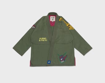 May include: An olive green martial arts gi jacket featuring yellow text that reads ALBINO & PRETO on the chest. The garment includes a yellow graphic on the shoulder, a small patch on the sleeve, and a colourful robot-like character patch.