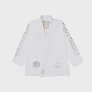 Op de afbeelding: Witte vechtsportuniform met gouden letters op de mouwen met de tekst "TOKYO ROLL". Het uniform heeft een wikkelontwerp met een bijpassende riem. Op de borst een ronde patch en de tekst "BORN X RAISER".