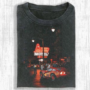 May include: Dark gray t-shirt featuring a graphic of a red race car with the number 95, in front of a neon sign that reads "Cozy Cone Motel". The image has a vintage, distressed look.