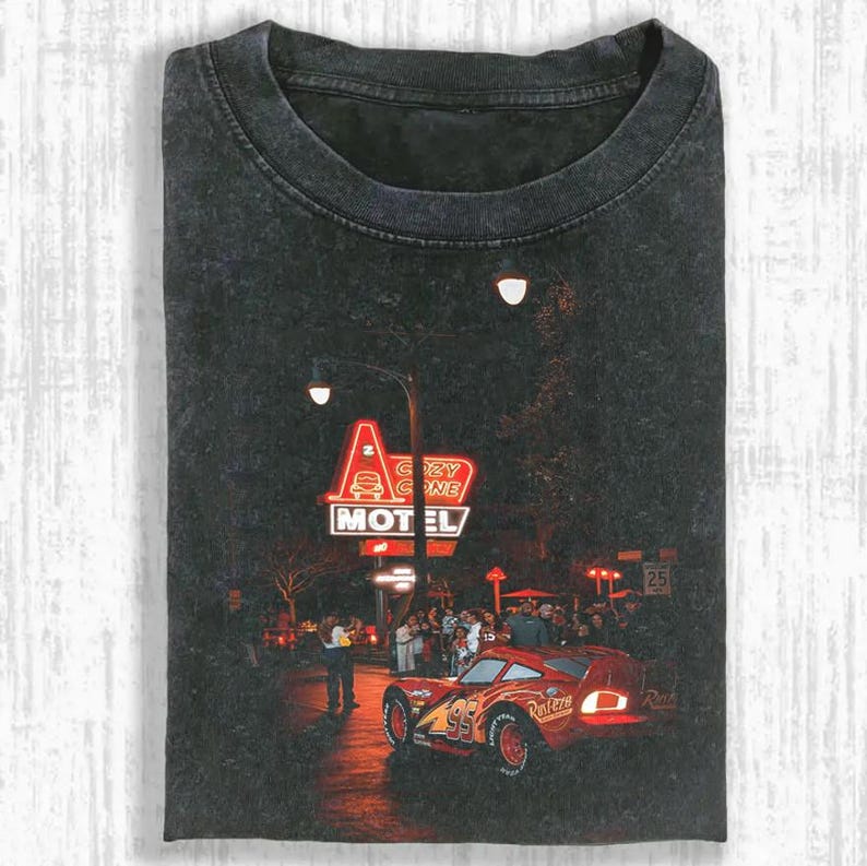 May include: Dark gray t-shirt featuring a graphic of a red race car with the number 95, in front of a neon sign that reads "Cozy Cone Motel". The image has a vintage, distressed look.