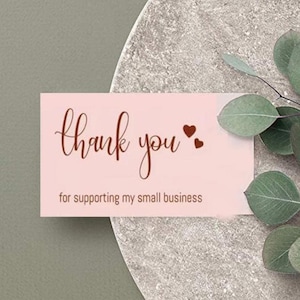 May include: A pink business card with the words "thank you" in a cursive font and two small heart shapes. The text "for supporting my small business" is below. The card is on a gray surface with green leaves.