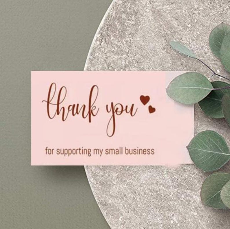 May include: A pink business card with the words "thank you" in a cursive font and two small heart shapes. The text "for supporting my small business" is below. The card is on a gray surface with green leaves.