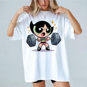 May include: A white short-sleeve t-shirt featuring a graphic print of a cartoon character with black hair and green eyes lifting a heavy grey barbell. The character wears a green top, pink shorts, and black shoes, surrounded by small yellow star accents.