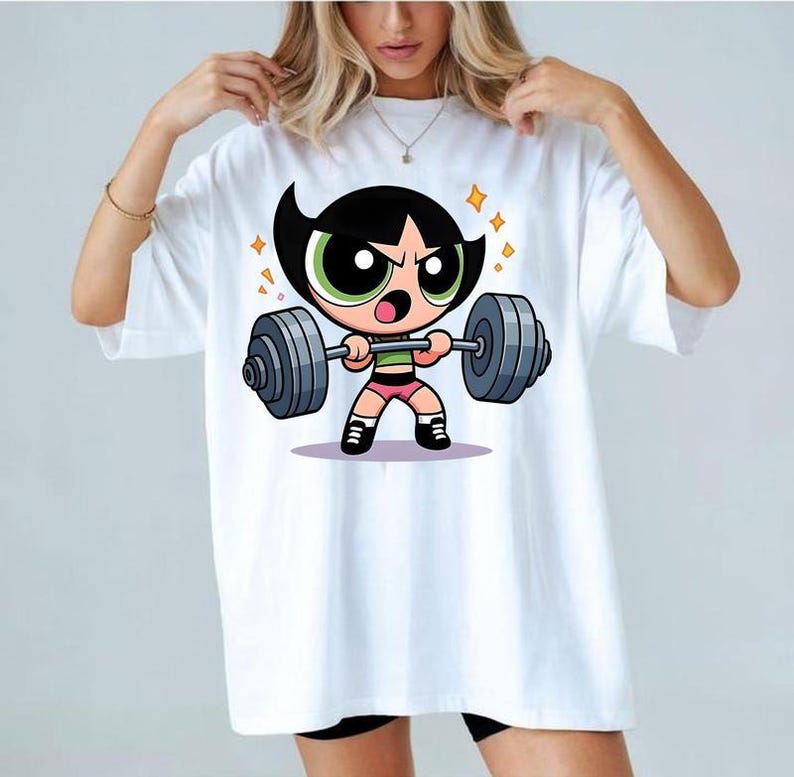 May include: A white short-sleeve t-shirt featuring a graphic print of a cartoon character with black hair and green eyes lifting a heavy grey barbell. The character wears a green top, pink shorts, and black shoes, surrounded by small yellow star accents.