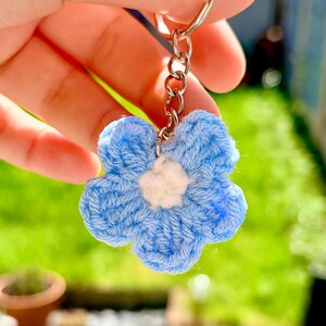 May include: A handmade blue and white crochet flower keyring. The flower has a light blue petal design with a white centre. The keyring is attached to a silver metal ring and chain.