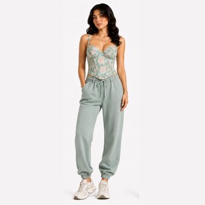 May include: A woman wearing a floral print corset top and light green sweatpants. The corset has a sweetheart neckline and thin straps. The sweatpants have a drawstring waist and elastic cuffs. The woman is wearing white sneakers.