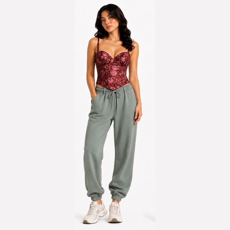 May include: A burgundy corset top with a floral pattern is paired with sage green sweatpants. The top has thin straps and a sweetheart neckline. The sweatpants have a drawstring waist and elastic cuffs. The model is wearing white sneakers.