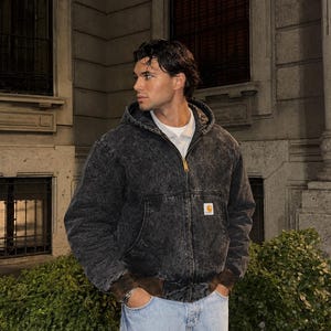 May include: A charcoal grey acid-wash hooded jacket with a front zip fastening, a white undershirt, and a small rectangular Carhartt logo patch on the front pocket. The jacket features dark brown ribbed cuffs and a matching brown ribbed hemline.