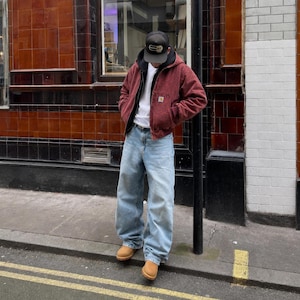 Puede incluir: Una chaqueta con capucha de color burdeos con cremallera, sobre una camiseta blanca lisa. El conjunto incluye unos vaqueros azul claro de corte holgado y botas de ante color beige. Una gorra de béisbol negra completa el atuendo casual.