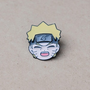 May include: A metal enamel pin featuring the head of a cartoon character with spiky yellow hair, a black headband with a grey metal plate displaying a spiral symbol, closed eyes, and an open mouth showing a pink tongue and white teeth.