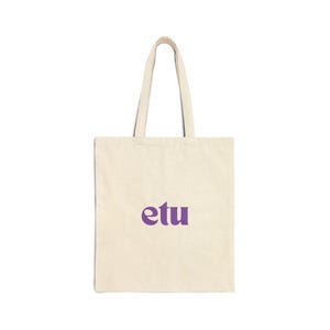 Peut inclure: Sac fourre-tout crème avec le mot "etu" en violet. Le sac est de forme rectangulaire avec une anse qui s'étend du haut. Il est fait d'un matériau semblable à de la toile.