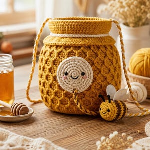 May include: A mustard yellow crochet bag shaped like a honey pot with a textured honeycomb pattern. The bag features a round cream-colored face applique with a smiling expression and a small attached crochet bee with white felt wings.