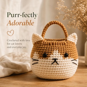 May include: A small, round, crocheted basket shaped like a cat head with a single handle. The basket features a tan and cream colour scheme, black plastic eyes, and embroidered black whiskers and a nose. Text reads: Purr-fectly Adorable.