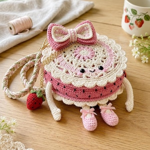 Pode incluir: Uma bolsa redonda de crochê em formato de torta de morango com corpo rosa, acabamento em renda creme e um laço grande no topo. Possui um rosto sorridente, braços e pernas pendentes com sapatos rosa, uma alça trançada e um pingente de morango.