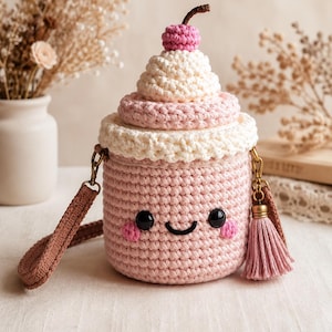 May include: A cylindrical crochet bag shaped like a milkshake, featuring a light pink body with a smiling face, black eyes, and pink cheeks. The top is cream-colored with a pink cherry on top, a brown strap, and a pink tassel attached to the side.