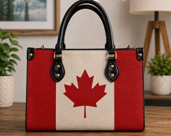 May include: A structured rectangular handbag featuring the Canadian flag design with a central white panel and a red maple leaf, flanked by red side panels. It has two black top handles, gold-tone metal hardware, and black trim along the edges.