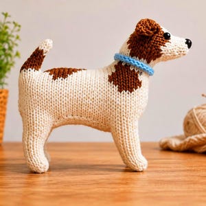 Könnte beinhalten: Ein handgestricktes Stofftier in Form eines Hundes mit cremefarbenem Körper und braunen Flecken auf Rücken, Kopf und Schwanz. Das Plüschtier hat schwarze Perlenaugen, eine kleine schwarze Nase und trägt ein hellblaues Strickhalsband.
