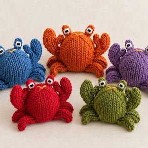 Könnte beinhalten: Fünf kleine, handgestrickte Krabbenspielzeuge in Blau, Orange, Violett, Rot und Grün. Jede Krabbe hat einen runden Körper, zwei erhöhte Augen mit schwarzen Pupillen und zwei große Scheren. Die Strickstruktur ist auf jeder Figur sichtbar.