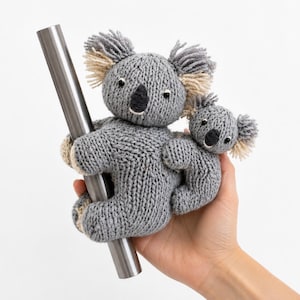 May include: A hand-knitted gray koala parent and baby toy set. The larger koala features textured gray yarn, cream-colored ear tufts, and a black nose, while the smaller koala clings to its side. Both are attached to a smooth, silver-colored metal rod.