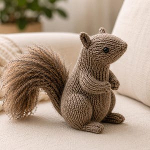 Könnte beinhalten: Ein braunes, gestricktes Eichhörnchen-Spielzeug mit einem strukturierten, flauschigen Schwanz und dunklen Kunststoffaugen. Der Körper ist aus einem glatten, gleichmäßigen Strickmuster gefertigt, während der große, geschwungene Schwanz eine ausgeprägte, fusselige und voluminöse Garnstruktur aufweist.