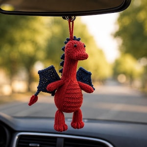 May include: A small, hand-knitted red dragon ornament with dark blue wings and a matching blue spine ridge. The creature features black bead eyes and is suspended by a red yarn loop, designed to hang from a vehicle rearview mirror.