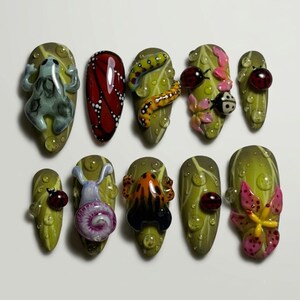 May include: A set of ten press-on nails featuring a nature-inspired theme with green leaf backgrounds and raised 3D accents including a gray frog, a red butterfly wing pattern, yellow caterpillars, ladybugs, a purple snail, an orange frog, and pink flowers.