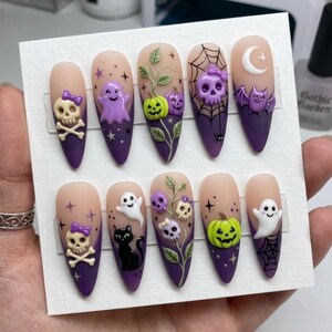 Könnte beinhalten: Ein Set aus zehn mandelförmigen Press-on-Nägeln mit violettem Farbverlauf, verziert mit 3D-Halloween-Anhängern und handgemalten Motiven wie Totenköpfen mit Schleifen, Geistern, Kürbissen, einer schwarzen Katze, Spinnennetzen, Fledermäusen und Sternen.
