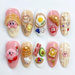 May include: A set of ten artificial nails with a breakfast theme. Each nail features a different design, including a croissant, fried egg, coffee, toast, and a cartoon character. The nails are painted in various colors, including pink, beige, and white.