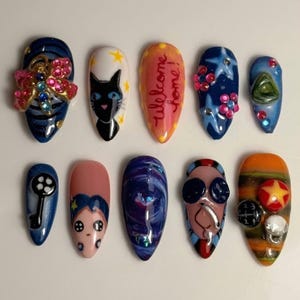 May include: A set of ten almond-shaped press-on nails featuring various designs including a pink rhinestone dragonfly, a black cat, the text "Welcome home!", blue stars, a black key, a character with button eyes, galaxy swirls, and colorful charms.
