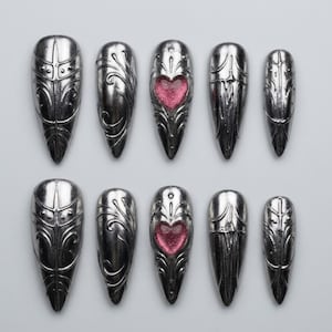 May include: A set of ten silver-toned press-on nails with a pointed almond shape. Each nail features intricate embossed metallic patterns, and the center nail in each row displays a recessed heart-shaped detail filled with sparkling pink glitter.