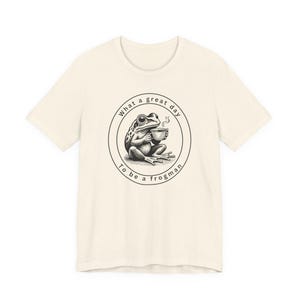 May include: Cream-colored t-shirt featuring a circular graphic of a frog holding a coffee cup. The graphic includes the text "What a great day to be a frogman" in a circular design. The shirt is made of a soft material.