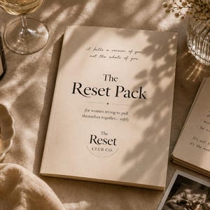 Op de afbeelding: Een brandende kaars in een zwart potje, een glas wijn en een boek met de titel "The Reset Pack" zijn gerangschikt op een beige oppervlak. Het boek bevat de tekst "voor vrouwen die proberen zichzelf bij elkaar te rapen... zachtjes". Gouden sieraden en een kleine vaas met bloemen zijn ook aanwezig.