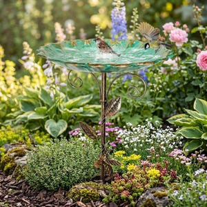 Puede incluir: Un bebedero decorativo con un cuenco de vidrio verde y un soporte de metal color bronce. Dos pájaros están en el bebedero, rodeados de un exuberante jardín con flores y vegetación coloridas. El bebedero tiene detalles de hojas y volutas.