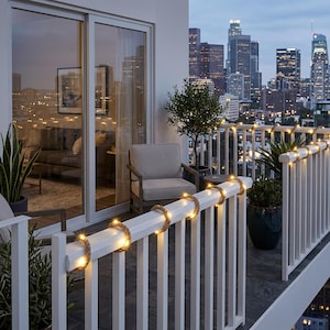 May include: A balcony with a white railing adorned with warm white string lights wrapped in rope. The balcony features a small tree, potted plants, and a chair. The background showcases a city skyline at dusk.