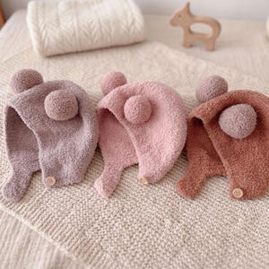 Peut inclure: Trois bonnets en polaire douce et duveteuse dans des tons violet cendré, rose clair et terre cuite. Chaque bonnet présente deux pompons ronds sur le dessus et une petite fermeture à bouton sur le rabat latéral, disposés sur un tissu texturé.