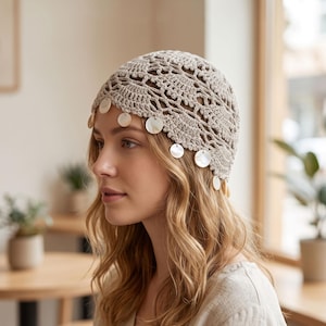 May include: A beige crochet beanie featuring an intricate openwork pattern and a scalloped edge. The lower border is adorned with small, round, iridescent white shell discs that hang freely from the crocheted fabric, creating a delicate texture.