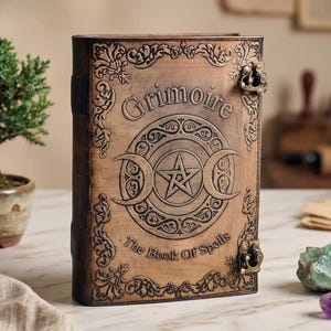 K&ouml;nnte beinhalten: Ein braunes, ledergebundenes Buch mit dem Wort "Grimoire" auf dem Einband. Der Einband zeigt ein zentrales Pentagramm-Design in einem Kreis, flankiert von Halbmonden. Der Text "The Book Of Spells" steht darunter. Zierliche Metallverschl&uuml;sse sichern das Buch.