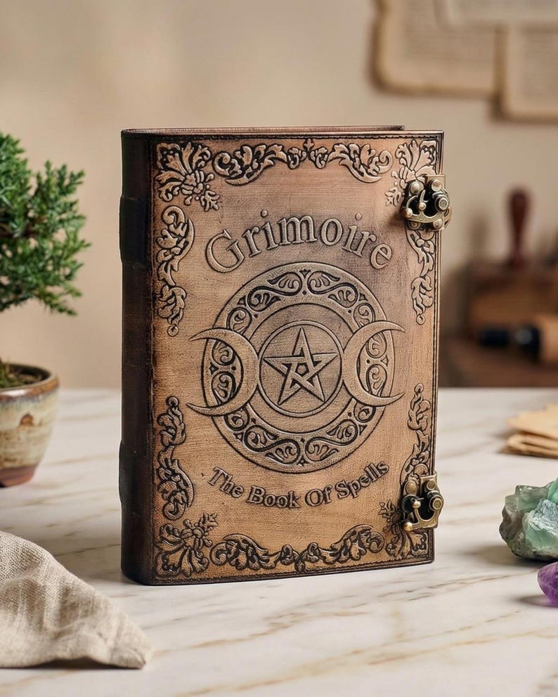 K&ouml;nnte beinhalten: Ein braunes, ledergebundenes Buch mit dem Wort "Grimoire" auf dem Einband. Der Einband zeigt ein zentrales Pentagramm-Design in einem Kreis, flankiert von Halbmonden. Der Text "The Book Of Spells" steht darunter. Zierliche Metallverschl&uuml;sse sichern das Buch.