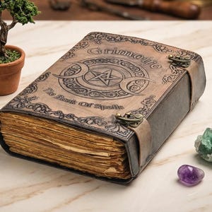 K&ouml;nnte beinhalten: Ein ledergebundenes Grimoire mit dem Titel "Grimoire" und "The Book Of Spells" auf dem Einband. Das Buch zeigt ein Pentagramm und ein Mondsichel-Design. Das Buch ist mit einem Lederriemen und Metallverschl&uuml;ssen gesichert. Ein kleiner Bonsai-Baum und Edelsteine befinden sich in der N&auml;he.