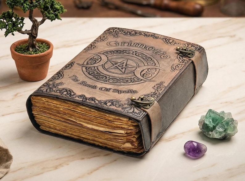 K&ouml;nnte beinhalten: Ein ledergebundenes Grimoire mit dem Titel "Grimoire" und "The Book Of Spells" auf dem Einband. Das Buch zeigt ein Pentagramm und ein Mondsichel-Design. Das Buch ist mit einem Lederriemen und Metallverschl&uuml;ssen gesichert. Ein kleiner Bonsai-Baum und Edelsteine befinden sich in der N&auml;he.